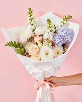Blush Blooms Bouquet featuring an assortment of light pink, green and blue flowers. The bouquet is wrapped in white tissue paper with a thin pink ribbon. The flowers are handheld against aa soft pink background.