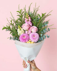 A small flower bouquet featuring two pink roses, two pink chrysanthemum and aa snap dragon as well as greenery. The bouquet is wrapped in white tissue paper and handheld on a pink background.